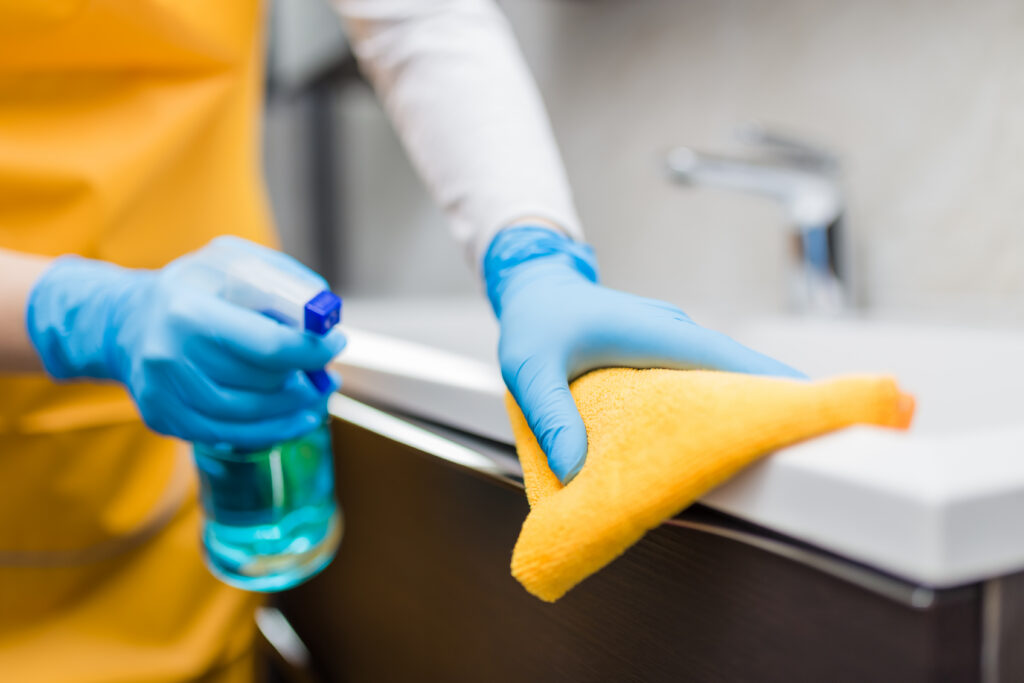 Commercial cleaning close up of female hands in rubber glover cleaning bathroom basin edge with sprayer disinfectant and microfiber cloth.